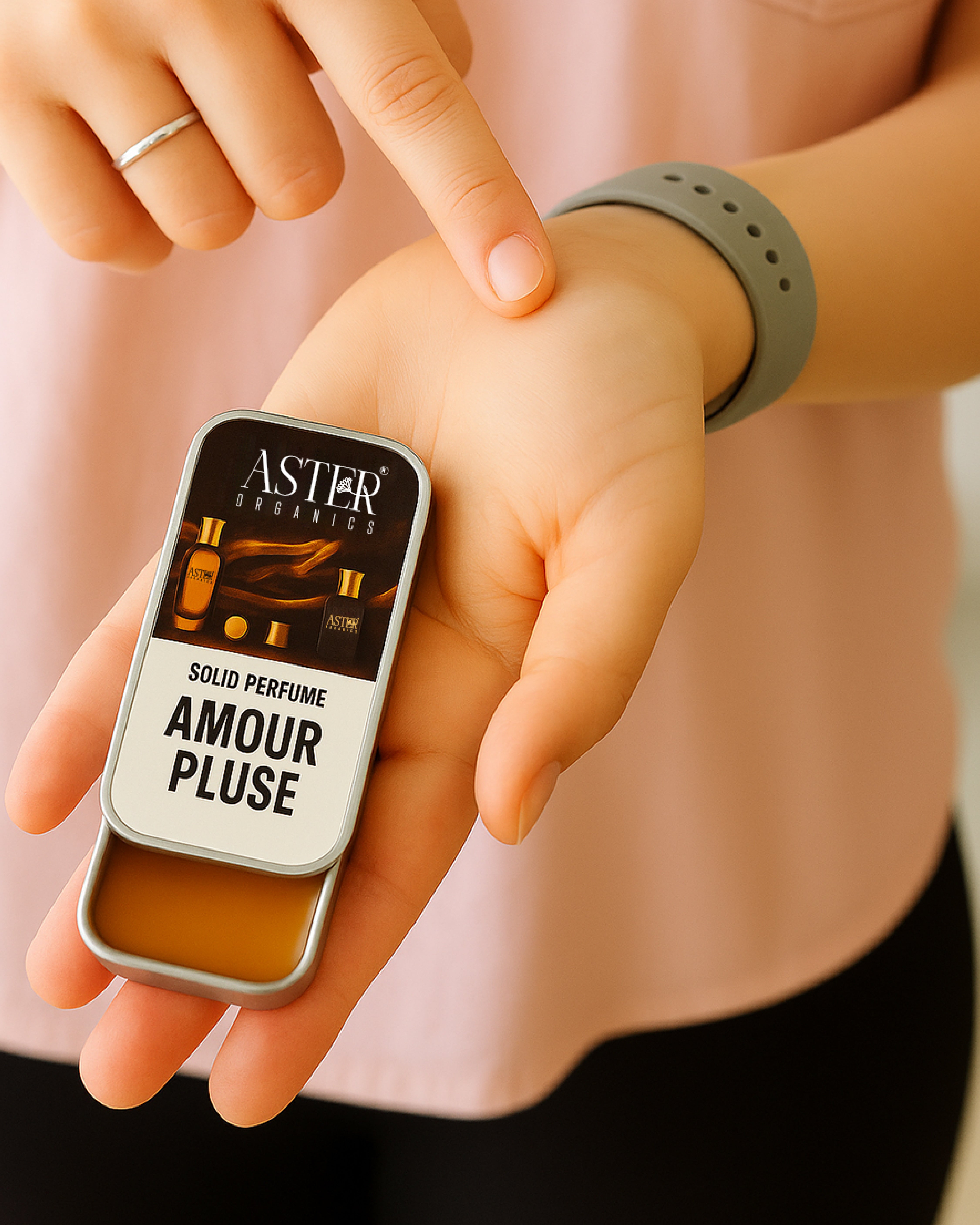 Solid perfume 'Amour Pluse' by Aster Cosmetics held between fingers with a blurred background.